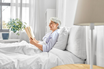 technology, old age and people concept - senior woman reading book in bed at home bedroom