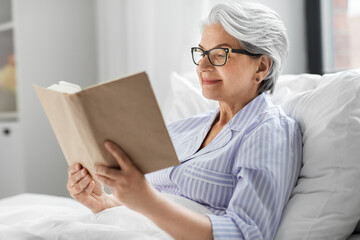 technology, old age and people concept - happy smiling senior woman in glasses reading book in bed...