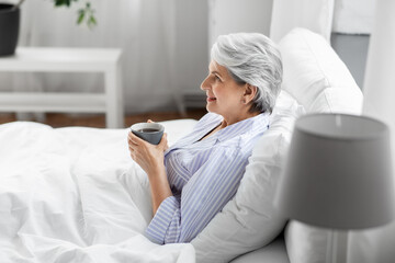 morning, old age and people concept - happy smiling senior woman in pajamas with cup of coffee sitting on bed at home bedroom