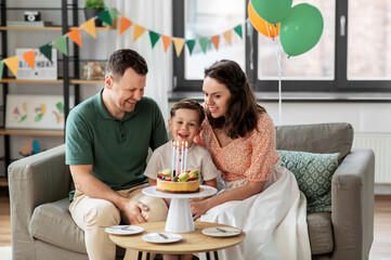family, holidays and people concept - portrait of happy mother, father and little son with four candles burning on birthday cake sitting on sofa at home party
