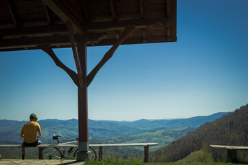 Fototapeta premium a cyclist dressed in yellow rests after an exhausting bicycle route on a bench on the edge of the mountain, admiring the beautiful views of the green mountains