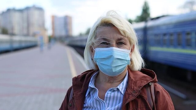 Close up senior woman with medicine mask on the face at public train station platform during coronavirus Covid-19 pandemic. Travel by train