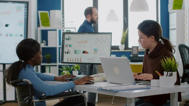African handicapped invalid entrepreneur in wheelchair working with project manager calculating profit on laptop, typing on pc, analysing financial documents sitting in start-up financial company.