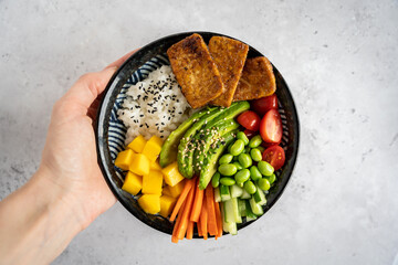 A female hand holding a homemade vegan poke bowl with tofu, mango, avocado on grey background