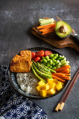 A homemade vegan poke bowl with tofu, mango, avocado, carrot, edamame, tomato, cucumber on black background