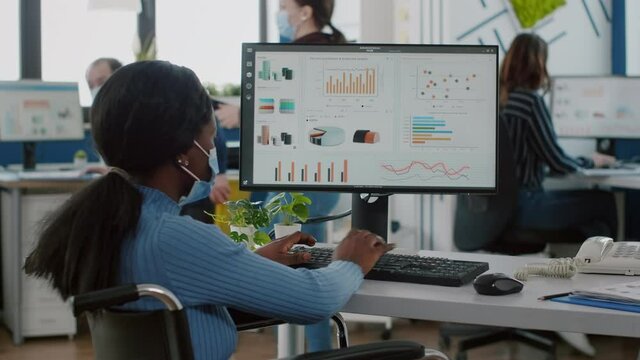 African invalid paralysed woman employee with protective mask immobilized in wheelchair working on computer checking reports, analysing datas from clipboard in strat up business financial office