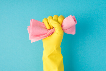 Photo of raised hand in yellow glove clenching pink rag into fist on isolated blue background with...