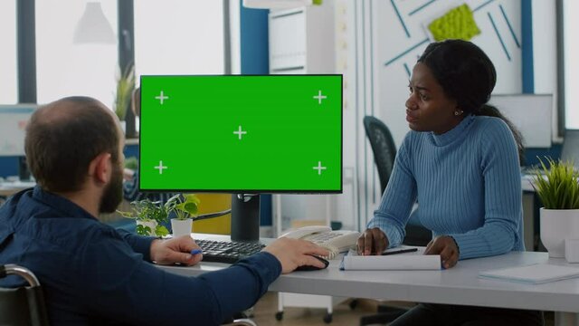 Handicapped paralysed entretreneur explaning to black employee financial evolution of company pointing at green screen, mock up, chroma key izolated desktop in start up office sitting in wheelchair
