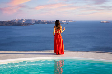 Beautiful woman in red dress  stands by the pool edge and enjoys the summer sunset with a glass of wine and view to the mediterranean sea