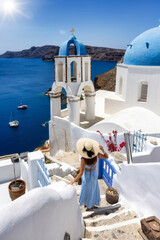 A elegant woman in a blue dress walks down next to the popular blue domed church in the little village of Oia, Santorini, Greece, during summer travel time