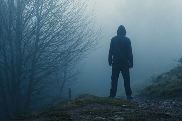 A scary hooded figure  on a path through a moody misty winter woodland. With trees silhouetted against the fog
