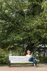 Fototapeta premium Woman resting in the park on bench and using mobile phone. Smiling lady chatting with someone in a messenger on a smartphone outdoors. Vertical photo