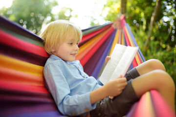Cute little blond caucasian boy reading book and having fun with multicolored hammock in backyard...