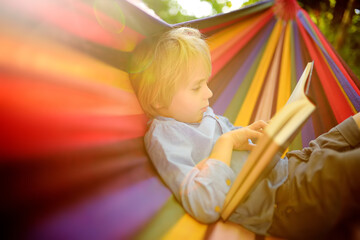Cute little blond caucasian boy reading book and having fun with multicolored hammock in backyard...