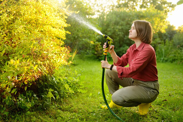 Young woman gardener watering plants with hose with sprinkler in sunny backyard. Gardening....