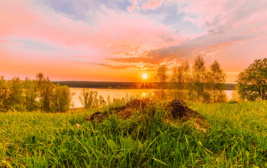 Obraz na płótnie Canvas Scenic view at beautiful sunset on a shiny lake with old rough stone on the foreground, green grass, birch trees, golden sun rays, calm water ,nice cloudy sky on a background, spring landscape