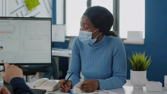 African employee with medical face mask explaning economic project to paralysed invalid handicapped coworker pesenting strategy of company. Multiethnical team working in startup financial office