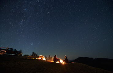 Young man and woman travelers sitting near bonfire under beautiful evening sky with stars....