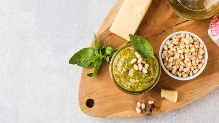 Pesto sauce in a jar with pine nuts, green basil and parmesan cheese on a wooden board. Top view. Copy space.
