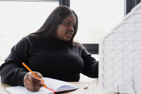 african american plus size designer writing in notebook while looking at house model.