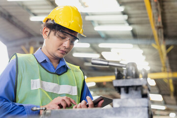 Manufacturing industry. Young male worker wearing hardhat and protection glasses with tablet look at machines in factory workshop.