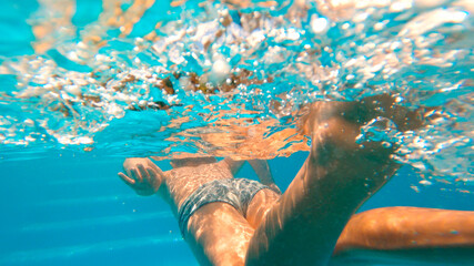 The body of Little kid swimming in a pool with swimsuit from the back, rear view. Underwater view. Beautiful shadows bubbles and reflections of the sun in the pool. Copy space.