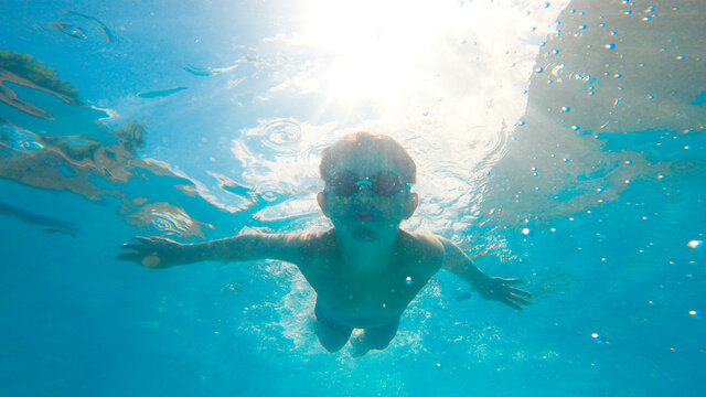 Underwater photo of a kid with goggles swimming into a pool with bubbles around and blue sky behind. Active healthy lifestyle, water sport activity and lessons on summer holidays vacation.