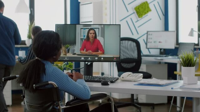 African disabled handicapped businesswoman sitting immobilized in wheelchair talking with remote partner on video call from startup business office. Black paralysed employee discussing project online