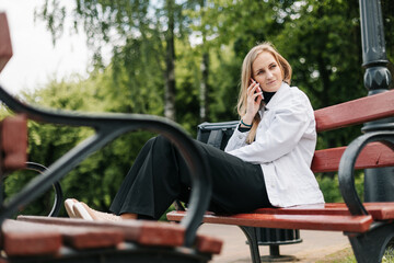 Happy blonde sitting on a bench in a city park, talking on the phone and texting with friends