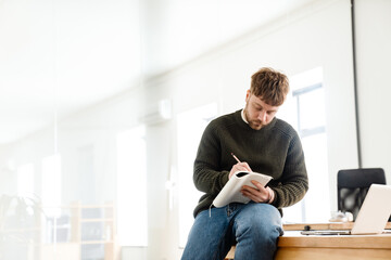 Young ginger man writing down notes while sitting on desk in office