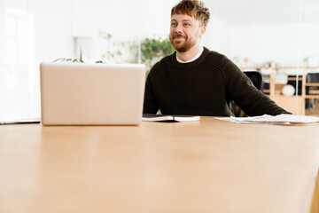 Young ginger man working with laptop while sitting at desk in office