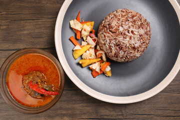Nasi dagang special brown white rice cooked with coconut milk pickled fruit vegetable pineapple carrot on black off white rim plate fish curry in bowl over rustic dark wooden background
