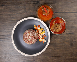 Nasi dagang special brown white rice cooked with coconut milk pickled fruit vegetable pineapple carrot on black offwhite rim plate chicken fish curry in bowl over rustic dark wooden background