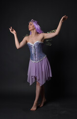 Naklejka premium Full length portrait of a purple haired girl wearing fantasy corset dress with fairy wings and flower crown. Standing pose against a dark studio background.