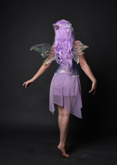 Full length portrait of a purple haired  girl wearing fantasy corset dress with fairy wings and flower crown.  Standing pose against a dark studio background.