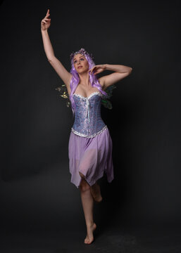Full length portrait of a purple haired  girl wearing fantasy corset dress with fairy wings and flower crown.  Standing pose against a dark studio background.