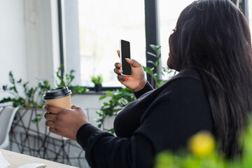 african american plus size businesswoman looking at smartphone with blank screen while holding paper cup.