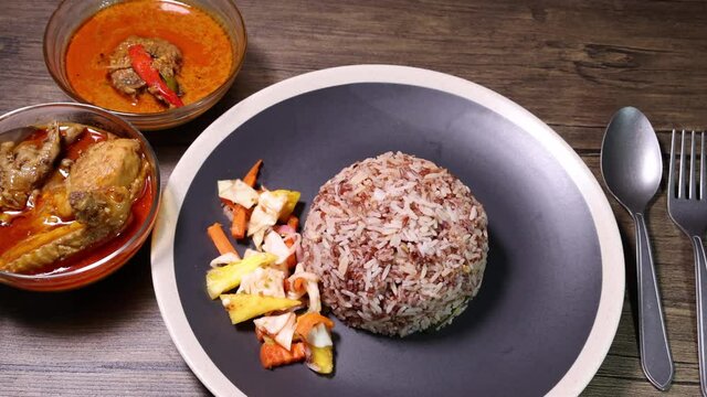 Nasi dagang special brown white rice cooked with coconut milk pickled fruit vegetable pineapple carrot black offwhite rim plate chicken fish curry bowl rustic dark wooden background fork spoon