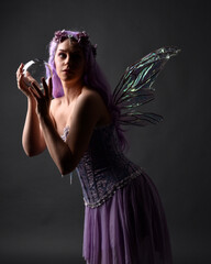 Close up portrait of a purple haired  girl wearing fantasy corset dress with fairy wings and flower crown, casting a spell.  Posing against a dark studio background with shadowed backlit lighting.