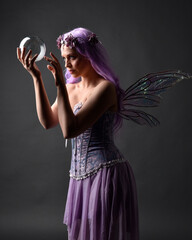 Close up portrait of a purple haired  girl wearing fantasy corset dress with fairy wings and flower crown, casting a spell.  Posing against a dark studio background with shadowed backlit lighting.