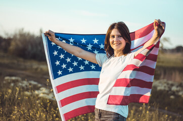 A beautiful young woman holding the national flag of America while standing in a field at sunset. 4th of July, Independence day. American freedom concept.