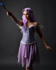 Close up portrait of a purple haired  girl wearing fantasy corset dress with fairy wings and flower crown, casting a spell.  Posing against a dark studio background with shadowed backlit lighting.