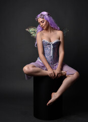 Full length portrait of a purple haired  girl wearing fantasy corset dress with fairy wings and flower crown.  Seated pose against a dark studio background.