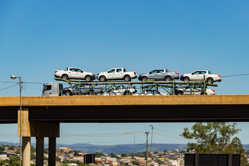 Um caminhão cegonha cheio de carros passando sobre um viaduto com céu azul ao fundo. - obrazy, fototapety, plakaty
