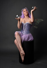 Full length portrait of a purple haired  girl wearing fantasy corset dress with fairy wings and flower crown.  Seated pose against a dark studio background.