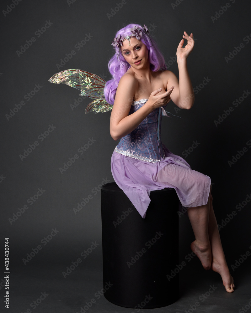 Poster Full length portrait of a purple haired  girl wearing fantasy corset dress with fairy wings and flower crown.  Seated pose against a dark studio background.