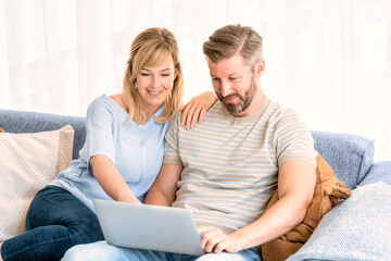 Smiling couple using laptop while sitting on the couch at home and browinsing on the internet