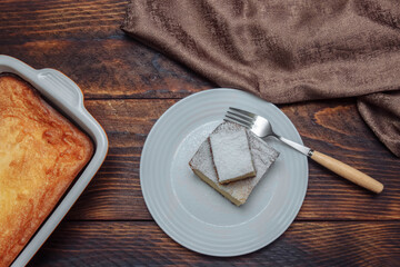 Cottage cheese casserole in baking dish and a slice in a white plate  with powdered sugar and fork on brown wooden background with napkin, top view, healthy breakfast concept, dessert cooking  recipe