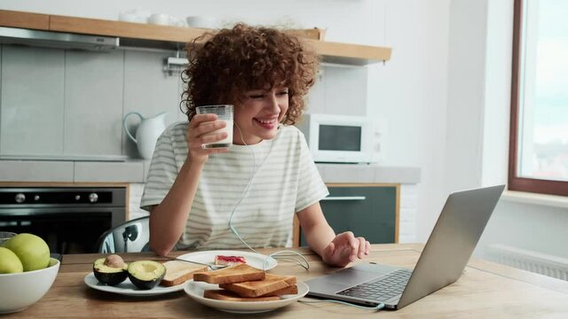 A Positive Curly-haired Girl In The Wired Headphone Talking At The Video Call On The Laptop And Eating Toast With Jam In The Kitchen