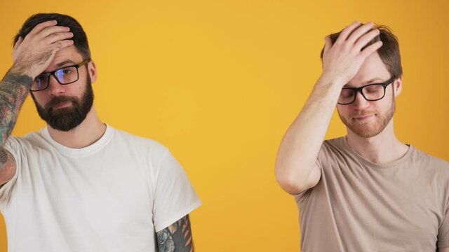 Two displeased guys wearing eyeglasses are doing face-palm standing isolated over yellow wall in the studio